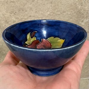 May include: A small, round ceramic bowl with a deep blue glazed finish. The interior features a hand-painted design of red fruit and green leaves. The bowl has a flared rim and a pedestal base, showcasing a glossy, reflective surface texture.