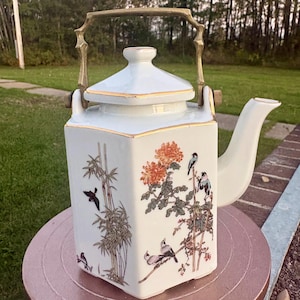 May include: A white porcelain teapot with a square body, a matching lid, and a gold-toned metal swing handle. The teapot features hand-painted illustrations of bamboo stalks, orange chrysanthemum flowers, and small birds on its exterior surfaces.