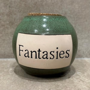 May include: A round ceramic jar with a speckled green glaze and a cork stopper. The front features a rectangular cream-coloured label with the word Fantasies printed in black serif font. The vessel has a wide body and a narrow opening at the top.