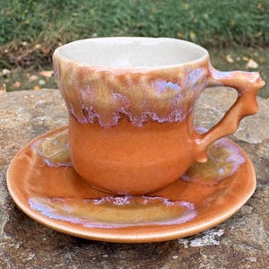 May include: A ceramic cup and saucer set featuring a vibrant orange base glaze with a thick, drippy cream and iridescent purple-tinted glaze cascading from the rim. The cup has a textured, organic-shaped handle and a smooth, off-white interior.