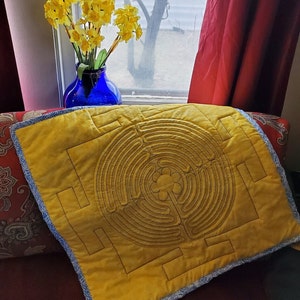 Puede incluir: Una almohada de tela amarilla con un diseño de laberinto bordado en marrón. La almohada está sentada en un sofá rojo con un patrón floral.