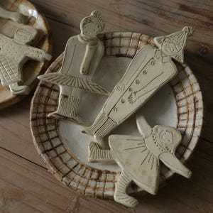 May include: A collection of off-white ceramic figures shaped like people, including a dancer in a tutu and a figure in a long coat, resting on two round, shallow ceramic plates with a brown grid pattern around the edges and a smooth centre.