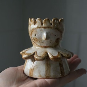 May include: A small, hand-sculpted ceramic egg cup featuring a stylized figure with a crown-like top, a pointed nose, and a wide, ruffled collar. The piece is finished in a glossy, off-white glaze with brown painted details and vertical stripes.