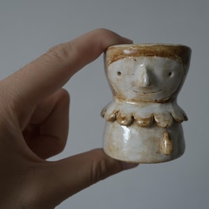 May include: A small, hand-sculpted ceramic cup featuring a stylized face with a pointed nose, simple dot eyes, and a thin line mouth. The piece has a scalloped collar detail and a glossy, off-white glaze with brown accents around the rim and base.