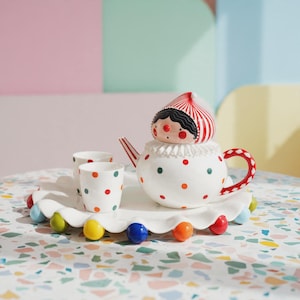 May include: A white ceramic tea set featuring a teapot with a clown-like lid, two small cups, and a scalloped tray. The pieces are decorated with colorful red, green, and orange polka dots. The teapot handle and lid have red and white stripes.