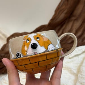 May include: A ceramic mug featuring a hand-painted illustration of a brown and white dog peeking over the rim. The lower portion of the mug is painted with a brown brick-like pattern, while the upper section is a light beige colour.
