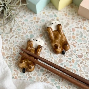 May include: Two ceramic chopstick rests shaped like brown and white sea otters lying on their backs. One otter rest supports a pair of dark brown wooden chopsticks, while the other rests nearby. Both figures feature small black eyes and noses.