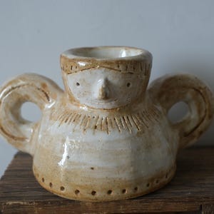 Può includere: Un vaso in ceramica fatto a mano caratterizzato da un volto umano stilizzato con dettagli incisi, un'ampia apertura circolare e due manici laterali prominenti. L'oggetto presenta una smaltatura color crema lucida con accenti marroni.