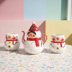 Peut inclure: Une théière en céramique blanche en forme de bonhomme de neige portant un chapeau rouge avec un sapin de Noël, accompagnée de deux tasses assorties. L'ensemble présente des écharpes rouges, des nez en carotte orange et des boutons colorés.