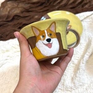 May include: A ceramic mug featuring a hand-painted illustration of a Corgi dog with brown, white, and tan fur. The mug has a glossy yellow exterior, a brown handle, and a matching yellow saucer visible in the background. The Corgi is depicted with its tongue out.