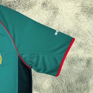 May include: Green soccer jersey with red trim and the Mexican national team emblem on the chest. The jersey has a dark green panel on the side and the word "ME" printed on the sleeve.