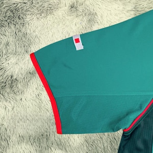 May include: Close-up of a teal athletic shirt sleeve with a red trim. The sleeve has a small white patch with a red square. The shirt has a mesh panel and a red accent line.
