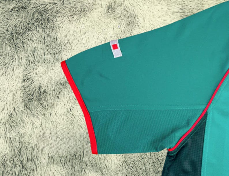 May include: Close-up of a teal athletic shirt sleeve with a red trim. The sleeve has a small white patch with a red square. The shirt has a mesh panel and a red accent line.