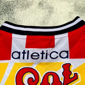 May include: Close-up of a red, white, and yellow athletic jersey. The collar is black and white striped. The word "atletica" is printed in navy blue, and the word "Col" is in red.