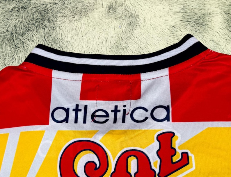 May include: Close-up of a red, white, and yellow athletic jersey. The collar is black and white striped. The word "atletica" is printed in navy blue, and the word "Col" is in red.