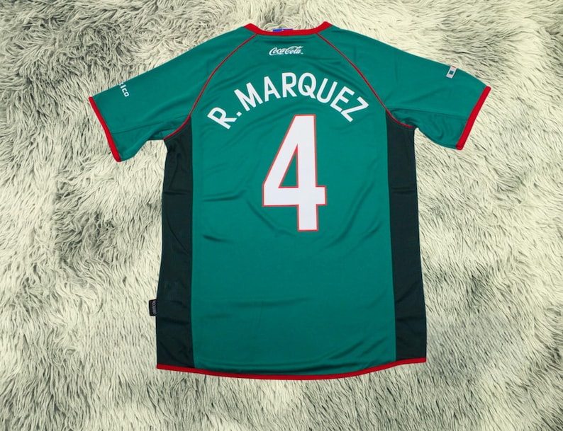 May include: A green soccer jersey with red trim, featuring the name "R. MARQUEZ" and the number "4" in white. The jersey has a Coca-Cola logo and a small emblem on the sleeve. The jersey is made of a breathable material.