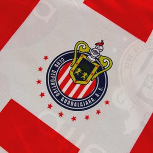 May include: Close-up of a white and red jersey with the Club Deportivo Guadalajara A.C. emblem. The emblem is a circular design with a shield, two lions, and the team's name. Red stars surround the emblem.