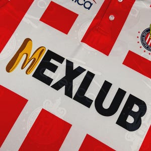May include: White and red soccer jersey with the word "MEXLUB" in black and gold lettering. The jersey has a pattern of red rectangles and a team logo on the upper right. The collar and button area are red.