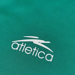 May include: Close-up of a teal fabric with the white word "atletica" and a stylized bird logo. The fabric has a subtle texture, and the logo is clean and modern. The overall aesthetic is sporty and athletic.