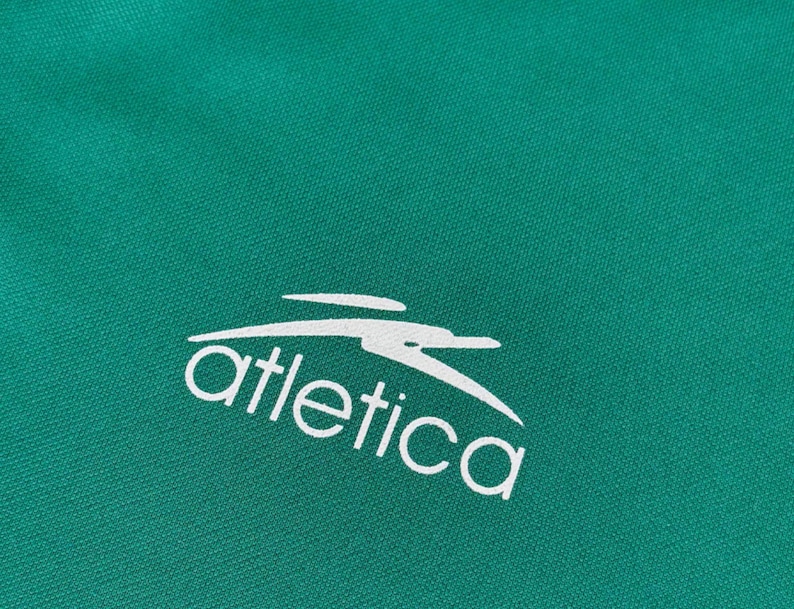 May include: Close-up of a teal fabric with the white word "atletica" and a stylized bird logo. The fabric has a subtle texture, and the logo is clean and modern. The overall aesthetic is sporty and athletic.