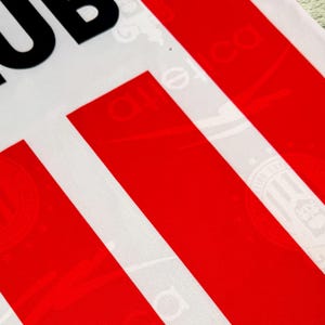 May include: Close-up of a white fabric with bold red stripes and the partial text "CLUB" in black. The fabric has a subtle pattern visible within the white areas, adding texture to the design. The overall aesthetic is clean and graphic.