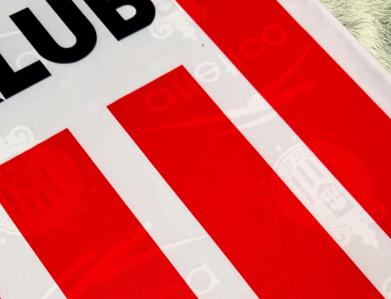 May include: Close-up of a white fabric with bold red stripes and the partial text "CLUB" in black. The fabric has a subtle pattern visible within the white areas, adding texture to the design. The overall aesthetic is clean and graphic.