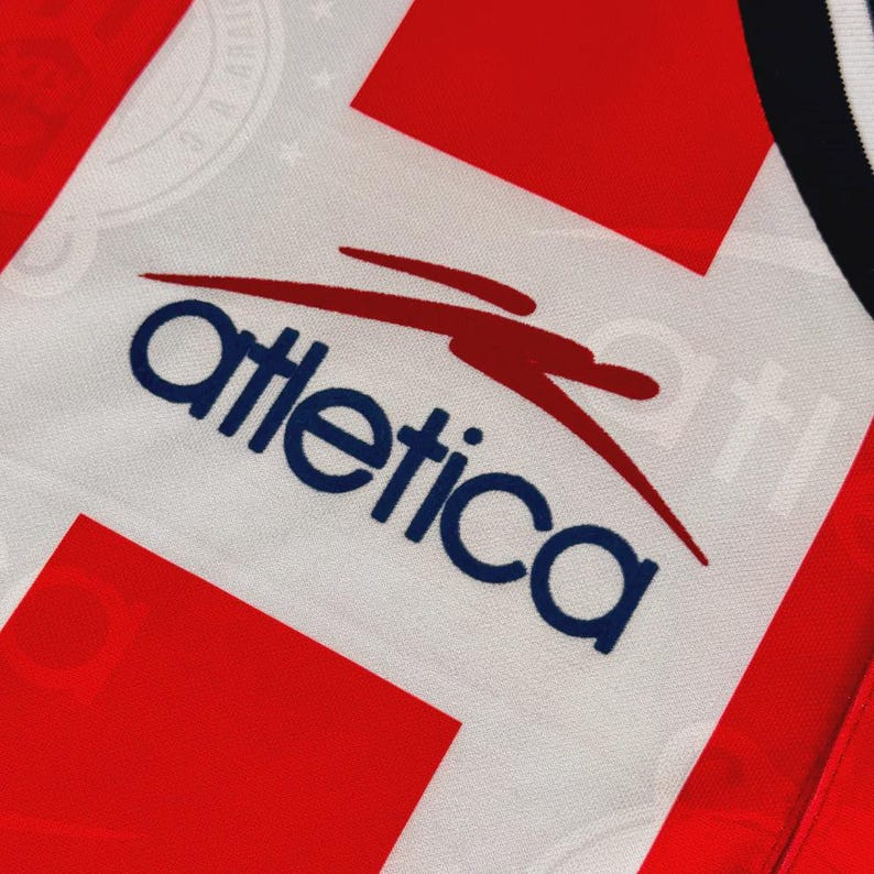 May include: Close-up of a white and red athletic jersey. The jersey features the word "Atletica" in navy blue with a red swoosh design. The jersey has a red and white pattern.