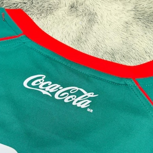 May include: Close-up of a teal athletic shirt with a red collar and trim. The Coca-Cola logo is printed in white on the shirt's front. The shirt is made of a textured, breathable fabric.