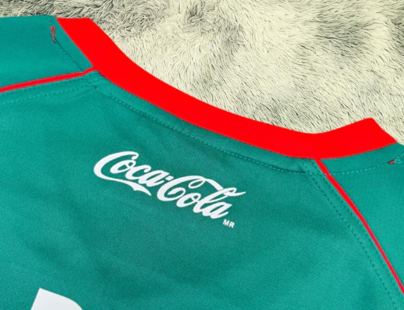 May include: Close-up of a teal athletic shirt with a red collar and trim. The Coca-Cola logo is printed in white on the shirt's front. The shirt is made of a textured, breathable fabric.