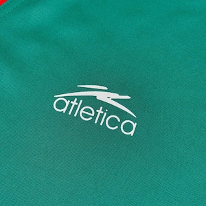 May include: Close-up of a green athletic jersey with a white logo and text that reads "atletica". The jersey has a red stripe at the top. The fabric has a textured appearance.