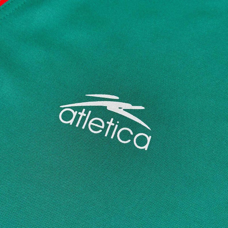 May include: Close-up of a green athletic jersey with a white logo and text that reads "atletica". The jersey has a red stripe at the top. The fabric has a textured appearance.