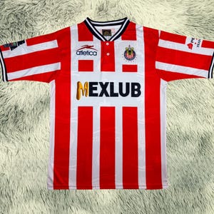 May include: A red and white striped soccer jersey with the word "MEXLUB" in gold lettering. The jersey has a white collar with black trim and the "Atletica" logo. The sleeves have red and white stripes with black trim.