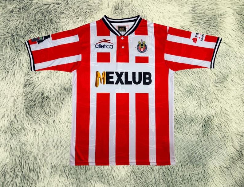 May include: A red and white striped soccer jersey with the word "MEXLUB" in gold lettering. The jersey has a white collar with black trim and the "Atletica" logo. The sleeves have red and white stripes with black trim.