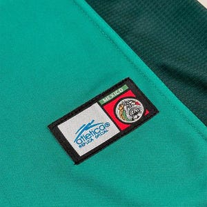 May include: Close-up of a green and dark green athletic jersey. A rectangular patch with the word "MEXICO" is at the top. Below the word "MEXICO" is a circular emblem with a white background and a blue logo. The words "Atletico Replica Oficial" are below the logo.