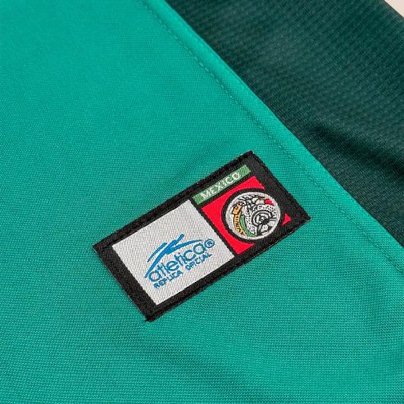 May include: Close-up of a green and dark green athletic jersey. A rectangular patch with the word "MEXICO" is at the top. Below the word "MEXICO" is a circular emblem with a white background and a blue logo. The words "Atletico Replica Oficial" are below the logo.