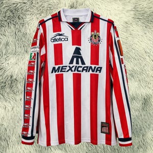 Może przedstawiać: Długi rękaw piłkarskiej koszulki z czerwono-białymi pionowymi paskami. Koszulka ma napis "MEXICANA" w kolorze granatowym, logo "Atletica" i herb drużyny. Rękawy mają powtarzający się wzór logo.