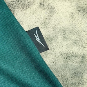 May include: Close-up of a teal fabric with a small black tag. The tag has a white graphic design. The fabric is textured and appears to be part of a garment. The background is a soft, light gray, fuzzy material.