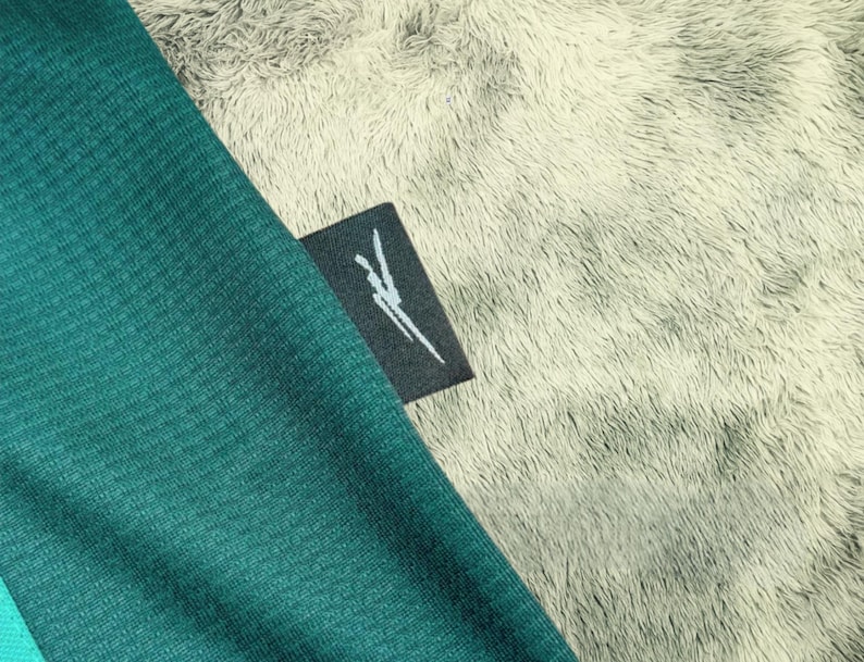 May include: Close-up of a teal fabric with a small black tag. The tag has a white graphic design. The fabric is textured and appears to be part of a garment. The background is a soft, light gray, fuzzy material.