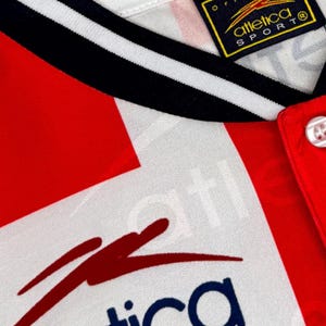 May include: Close-up of a vintage athletic jersey. The jersey features a red and white color scheme with black and white striped trim. The collar is white with black stripes. The jersey has a button-up front and a logo on the tag.