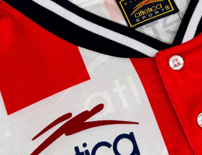 May include: Close-up of a vintage athletic jersey. The jersey features a red and white color scheme with black and white striped trim. The collar is white with black stripes. The jersey has a button-up front and a logo on the tag.