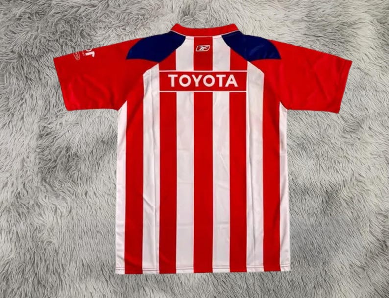 以下が含まれることがあります： 背中に「TOYOTA」の文字が入った赤と白のストライプのサッカーユニフォーム。ユニフォームは赤い袖と肩と襟に青いアクセントが付いています。襟にはReebokのロゴが見えます。