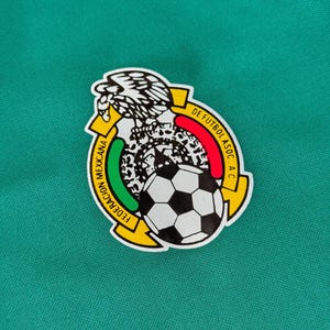 May include: A circular emblem featuring the Mexican Football Federation logo. The emblem includes a soccer ball, an eagle, and text in Spanish. The background is a solid teal green.