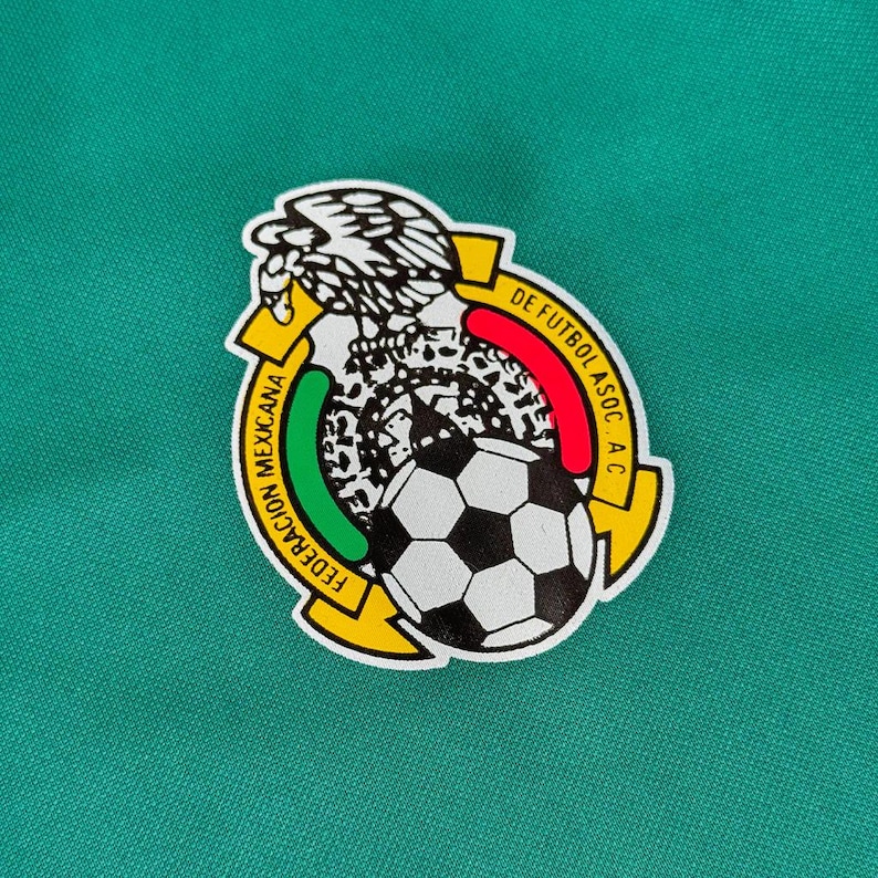 May include: A circular emblem featuring the Mexican Football Federation logo. The emblem includes a soccer ball, an eagle, and text in Spanish. The background is a solid teal green.