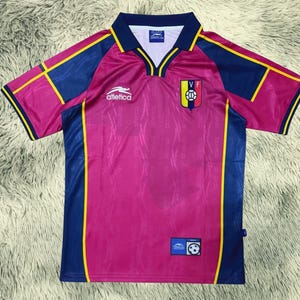 Peut inclure: Un maillot de football rose avec des détails bleus et jaunes. Le maillot a un col en V et des manches courtes. L'avant présente un écusson d'équipe et le mot "Atletica". Une petite étiquette avec un ballon de football est en bas.