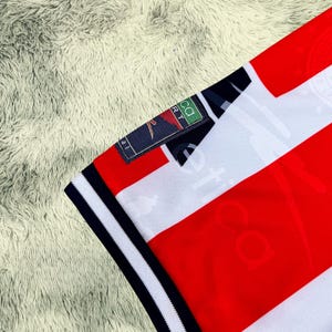 May include: Close-up of a red and white striped jersey with a navy blue collar and sleeve trim. The jersey has a small tag with the text "CA SPORT". The jersey is made of a soft, breathable material.