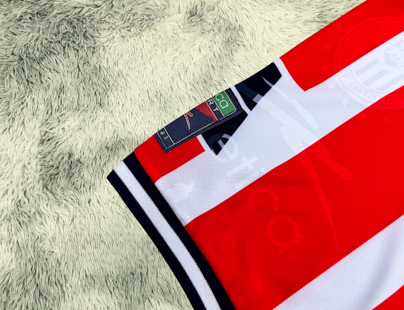 May include: Close-up of a red and white striped jersey with a navy blue collar and sleeve trim. The jersey has a small tag with the text "CA SPORT". The jersey is made of a soft, breathable material.