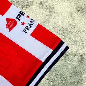 May include: Close-up of a white and red striped jersey with the text "PE FRAN" and a logo. The jersey has a navy blue and white striped trim on the sleeve. The jersey is on a fluffy, light-colored surface.