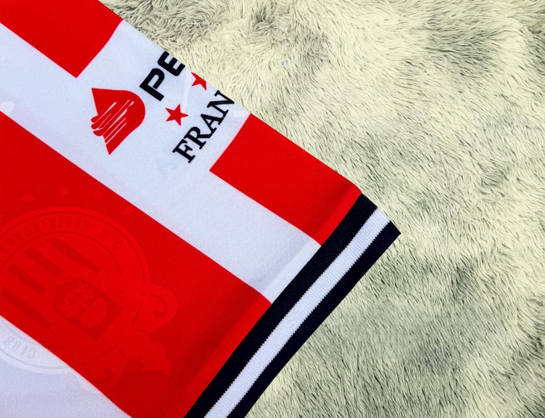 May include: Close-up of a white and red striped jersey with the text "PE FRAN" and a logo. The jersey has a navy blue and white striped trim on the sleeve. The jersey is on a fluffy, light-colored surface.