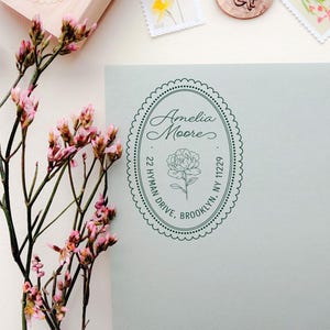 Puede incluir: Una vista aérea de un sobre gris claro con un sello de dirección personalizado y varios sellos florales. También se ven una caja de sellos de madera y dos sellos de lacre. El sello de dirección dice "Amelia Moore, 22 Hyman Drive, Brooklyn, NY 11229".