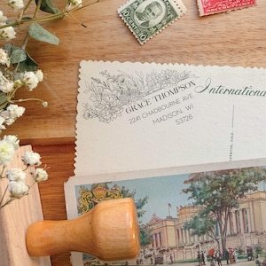 May include: A collection of vintage postcards and stamps, including a postcard with the text "International" and "Post Card". The stamps are red, orange, and green. A wooden stamp and sprigs of greenery are also present.